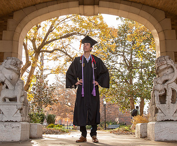 Student wearing cap and gown standing between lion statues under journalism archway
