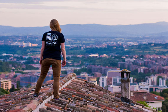 Student wearing t-shirt that reads "Take the world by the horns. Study abroad" standing on rooftop looking out over village